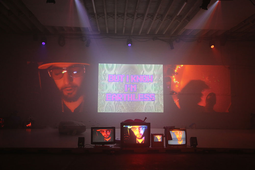 Alex Romania's Face Eaters at The Chocolate Factory Theater, Spring 2024. Photo by Brian Rogers. Image description: triptych of large video images, featuring a slowly rapping man wearing dark sunglasses on the left; pink text reading "But I Know I'm Earthless" at center; and a wobbly campfire at right. In the foreground, 4 small video monitors display similar images. Onstage: Alex Romania encased in a foam mattress wrapped in duct tape.