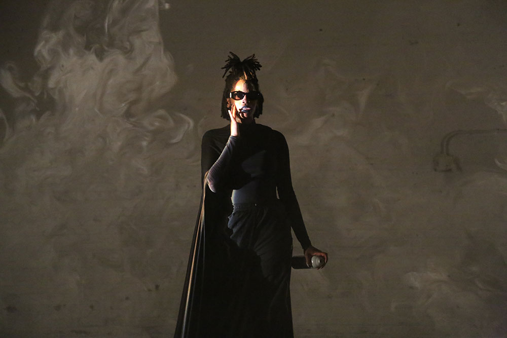 image from Angie Pittman's Black Life Chord Changes. Photo by Brian Rogers. Angie Pittman, wearing dark sunglasses and a large black cape, stands in the foreground. Haze billows around her.