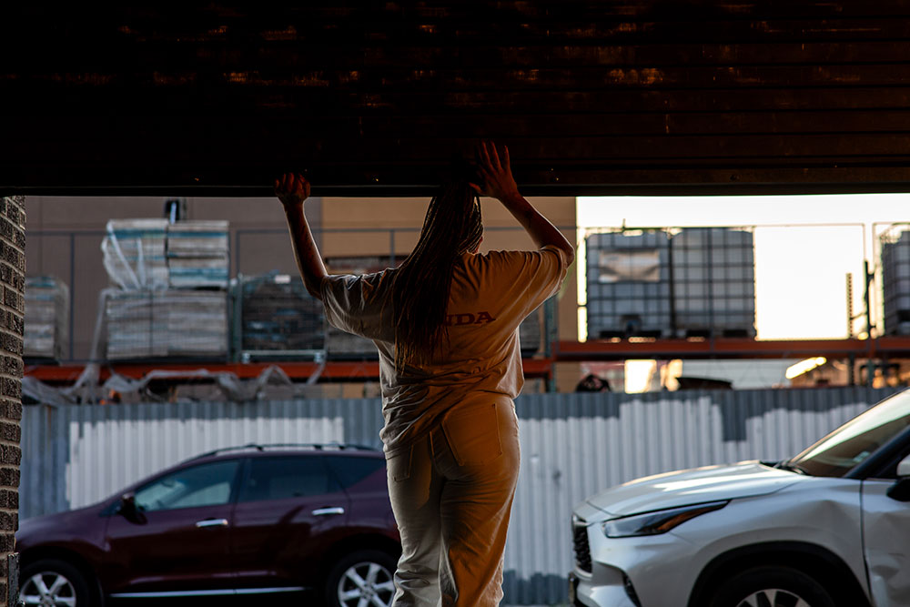 Image from jess pretty's "call and response". Photo by Rachel Keane. jess pretty, her back to us, wears a white top and white pants. She stands at the large steel roll gate, the entrance to The Chocolate Factory Theater. The roll gate has been opened to the street. jess's hands rest on the gate. In the background, parked cars, buildings, skyline.