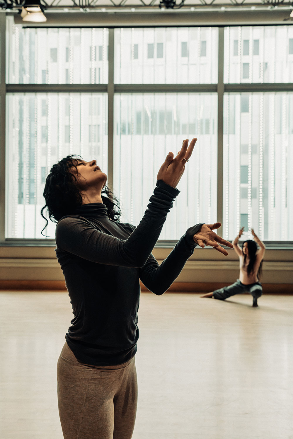 rehearsal photo (by Matt Vega) of Ayano Elson's Control. A rehearsal studio with a wall of windows and blond wood floor. In the foreground, Amelia Heintzelman, wearing a dark top and tan bottom, stands looking upward, one hand flatly extended, the other perpendicular. Behind, a performer crouches, arms extended.