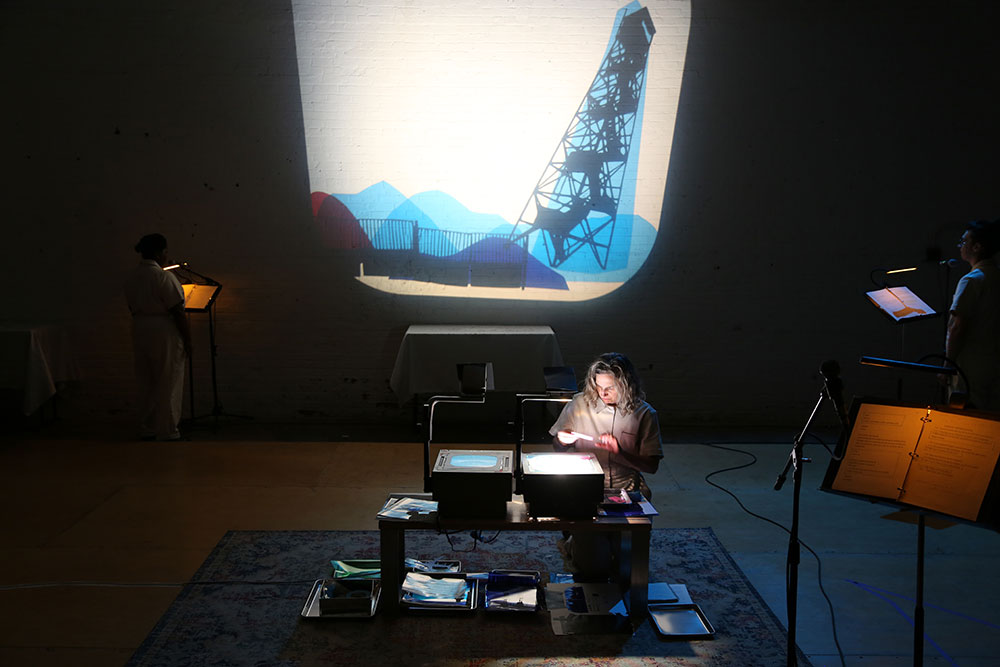 image from Your Ghost Body by Karinne Keithley Syers. Sarah Walsh kneels behind two overhead projectors. She manipulates a series of transparencies. Behind, the image of blue mountains and a leaning tower. To the right, a dimly lit music stand.
