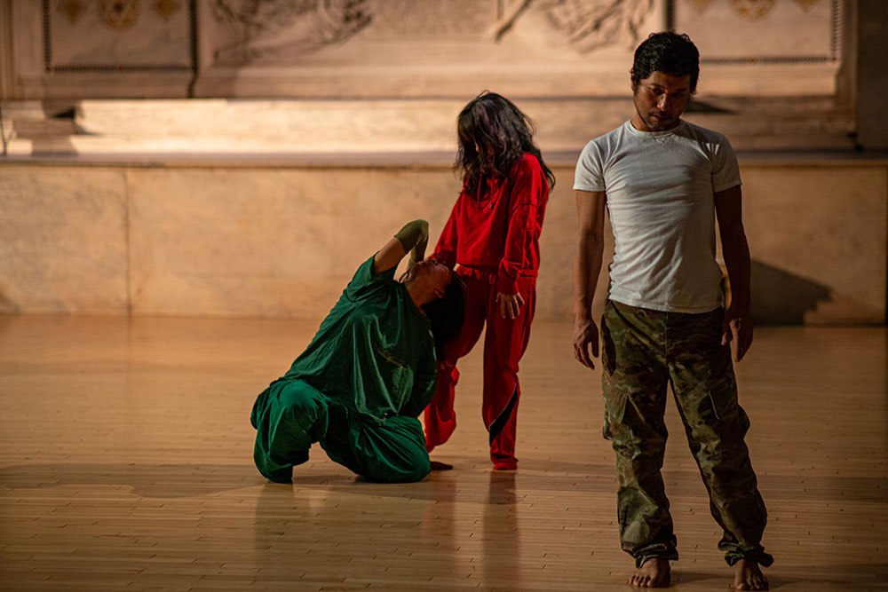 image by Rachel Keane from lu yim's Bottom Edge Air, performed at Judson Church. Three performers are positioned before the church altar. One, wearing green, crouches beneath a second performer dressed entirely in red. In the foreground, a third performer, wearing a white shirt and pants in a camouflage pattern, looks down.