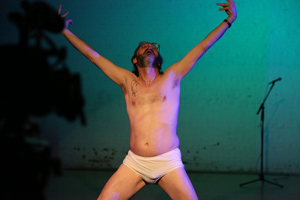 Neal Medlyn, in white underwear, stands with legs and arms spread wide, looking to the sky. In the background, a white wall lit in a blue wash, and a microphone stand.