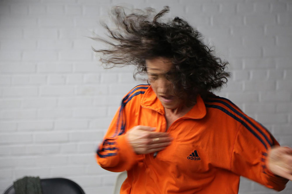 blurry photograph of Netta Yerushalmy at the dress rehearsal of "nothing personal, just everything". Netta, wearing a striped orange top, is pictured before a white brick background.