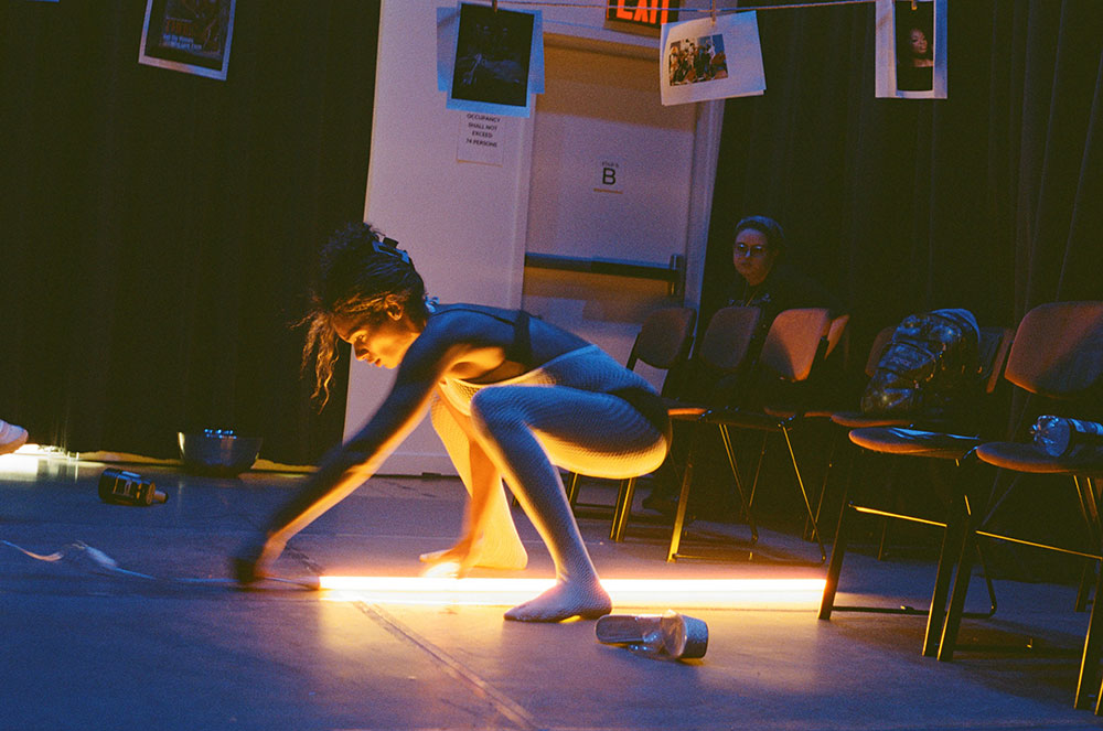 rehearsal image by Clifford Prince King of Zerina Tye's Glossolalia. A performer wearing a mesh onesie straddles a fluorescent light on a marley-covered floor. To the right, a row of chairs. Above, photos are mounted to a long string with clothespins.