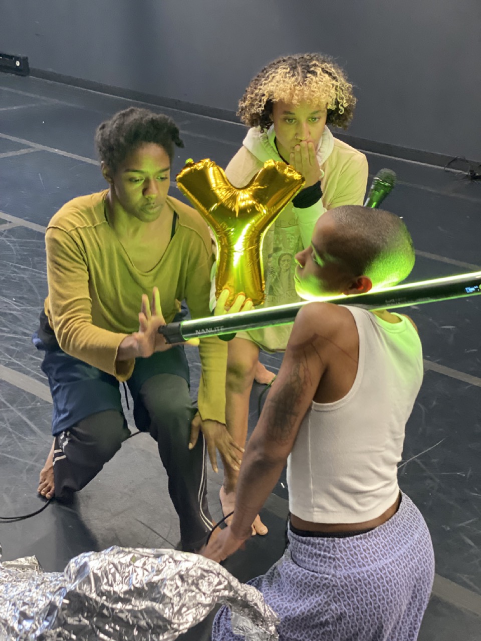 rehearsal image from Autumn Knight's Nothing: More. Kaylah Farrish, Dominica Greene, Jasmine Hearn, each wearing rehearsal clothing, kneel on a marley-covered floor. Jasmine cradles an LED light above her shoulder.