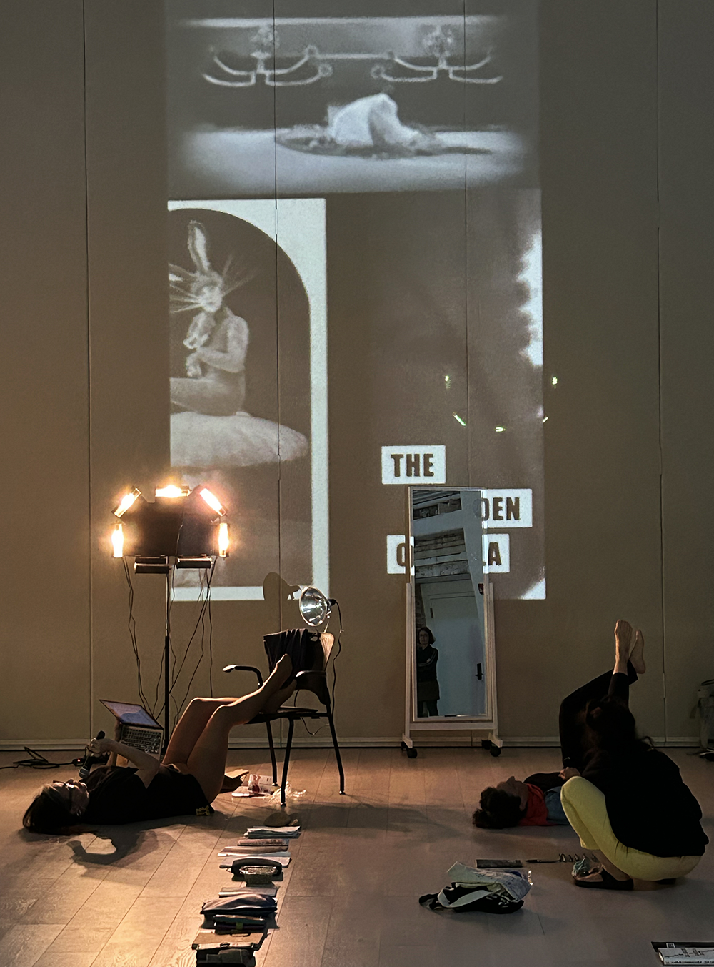 rehearsal image from "nothing personal, just everything" by Mieke Ulfig. In a studio with white walls and wood floors, a black and white video is projected on the rear wall. A long mirror leans against the wall, next to a light sculpture. Objects are arranged on the floor in neat rows. One performer lays on the floor, her legs resting on a chair. Another kneels, facing toward the wall.