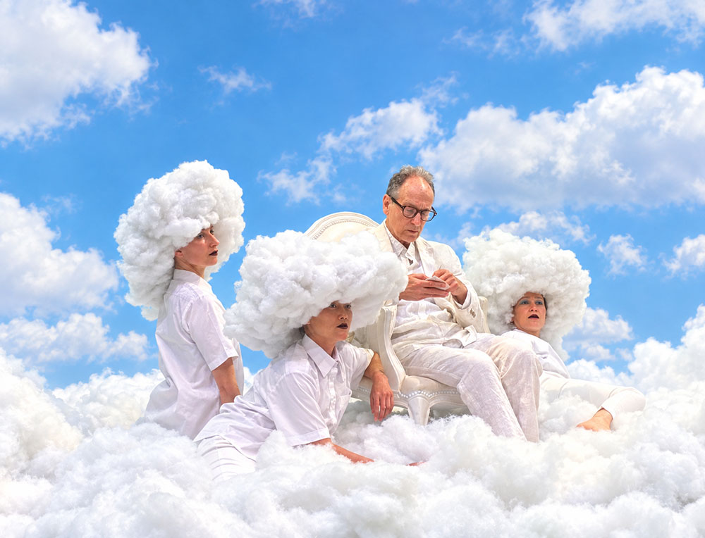 Image (by Annie-B Parson) from a rehearsal of "Lecture On The History Of Skywriting" at MIT in Summer 2026. Before a vivid blue background, Paul Lazar sits atop a bed of fluffy clouds. Sarah Konner, Caitlin Canty, and Cassie Wang, clouds perched atop their heads, kneel nearby.