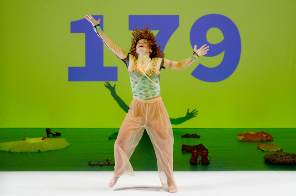 1000_liz_pierre_2026 performance still from This Is Unreal by Liz Santoro + Pierre Godard. In the background, a green screen projection with the numbers 179 in large blue font at the center, and a collection of dance-related objects situated on a green marley floor. Liz stands in the foreground, dressed in loose-fitting dance clothes, her gaze lifted upwards and her arms wide.