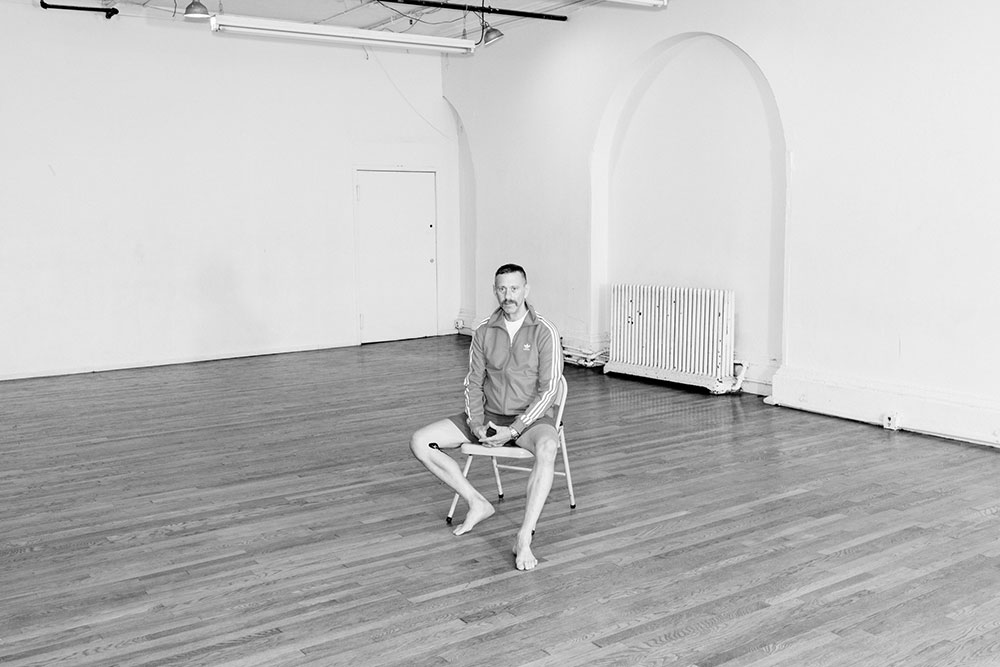 1000_neilgreenberg Black and white photo of Neil Greenberg by Mark Sommerfeld. Neil sits on a folding chair in the center of wood-floored rehearsal studio. Behind him, a radiator is nestled within an archway. Neil, wearing shorts and a striped exercise jacket, look toward the camera.