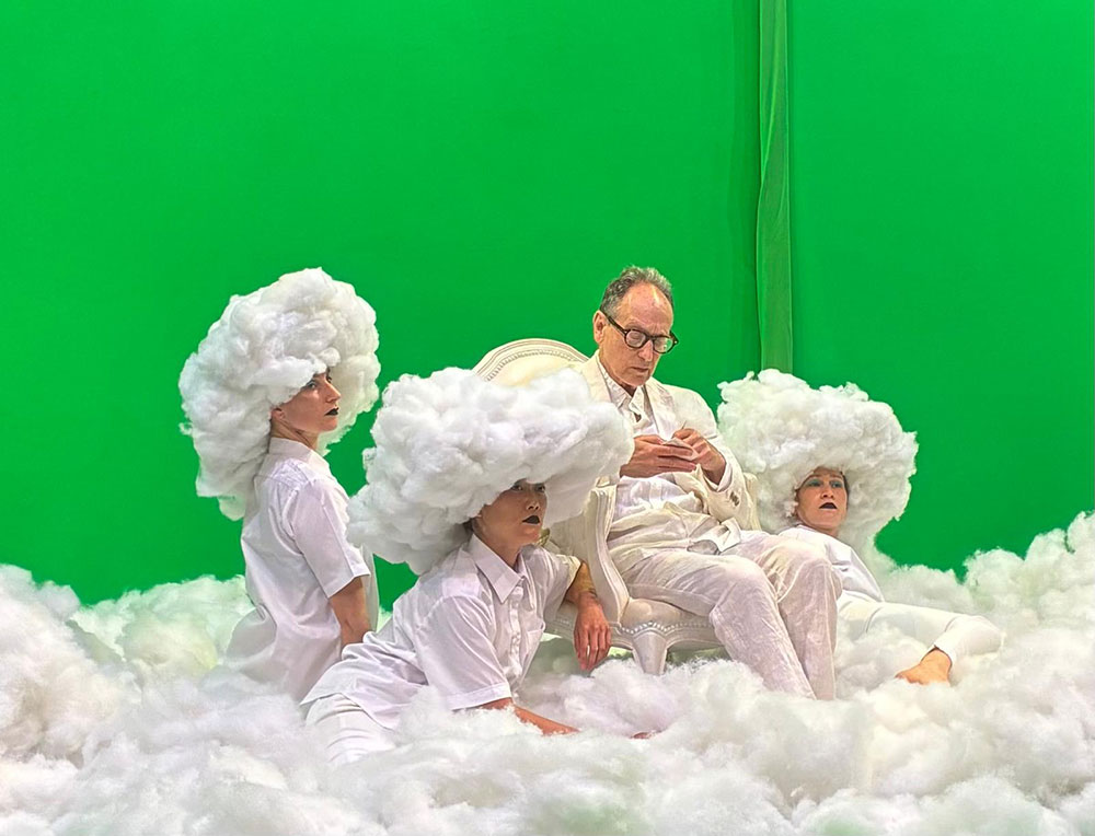 Image (by Annie-B Parson) from a rehearsal of "A Lecture On Skywriting" at MIT in Summer 2026. Before a vivid green screen background, Paul Lazar sits atop a bed of fluffy clouds. Sarah Kooner, Caitlin Canty, and Cassie Wang, clouds perched atop their heads, kneel nearby.