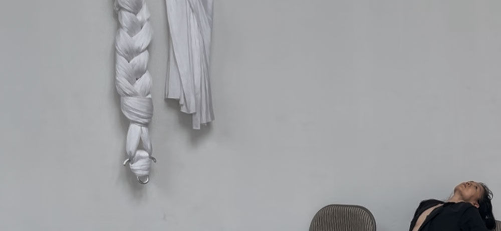 rehearsal image (by Sam Kim) from Kid Crone. A white aerial silk is draped against a flat white wall. Below, Sam sits on a folding chair, leaning back.