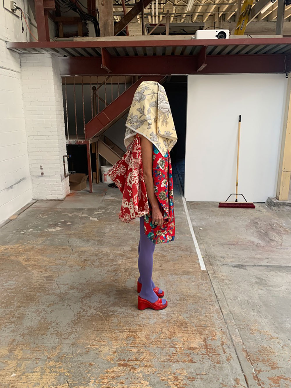 image from a performance by Walter Dundervill during an AUNTS event at The Chocolate Factory Theater in 2022. In the foreground of The Chocolate Factory's raw industrial space, a performer stands facing right, wearing a red patterned blouse, purple stockings and red shoes. A beige patterned cloth covers their head and face.